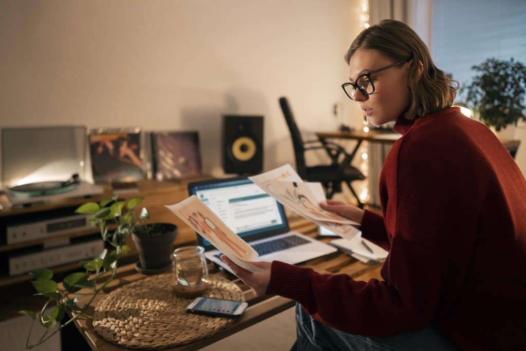 Uma mulher com óculos e suéter vermelho analisa ilustrações impressas em uma sala acolhedora com luz suave. Na mesa de madeira, há um notebook, uma planta, um copo de chá, e um celular. Ao fundo, um toca-discos, discos de vinil e uma cadeira de escritório compõem o ambiente criativo.