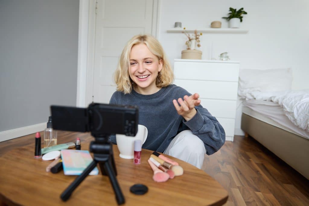 Uma jovem loira sorridente está gravando um vlog em seu quarto. Ela está sentada no chão com uma mesa baixa à sua frente, que contém pincéis de maquiagem, batons e outros produtos de beleza. Uma câmera em um tripé está posicionada para filmá-la, enquanto o quarto ao fundo está decorado de forma minimalista com móveis brancos e plantas.