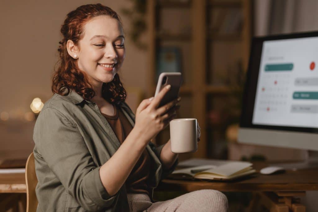 Mulher sorridente sentada à mesa segurando um celular e uma xícara de café. No fundo, um computador exibe uma tela com um calendário e notificações. O ambiente é aconchegante e iluminado com luz amarelada.