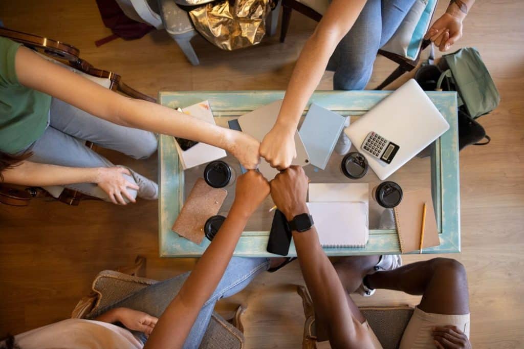 A imagem mostra um grupo de pessoas interagindo de forma colaborativa em um ambiente de trabalho. As mãos delas estão unidas em um gesto de "fist bump" (cumprimento com os punhos), simbolizando união e trabalho em equipe. No fundo, há uma mesa de trabalho com itens como computadores, cadernos e copos, o que sugere um ambiente focado em produtividade e comunicação.