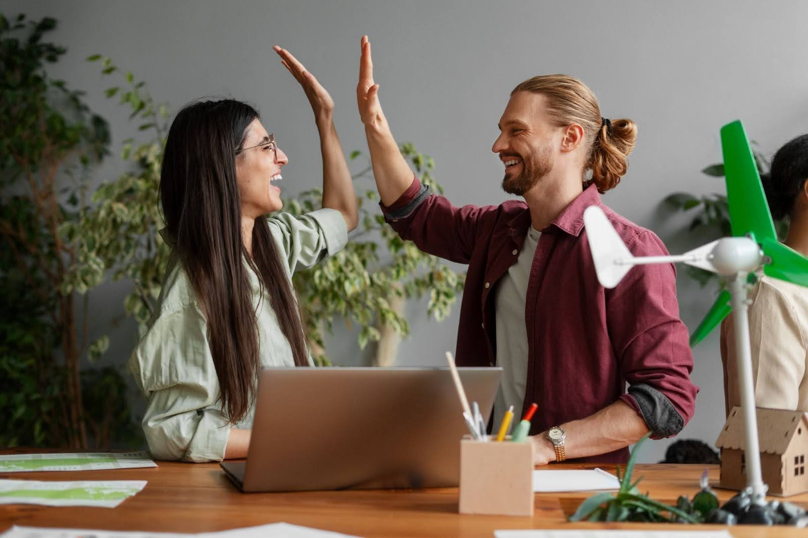A imagem mostra dois profissionais sorrindo e trocando um "high five" em um ambiente de trabalho. Eles estão ao lado de uma mesa de escritório, com um laptop aberto e elementos que sugerem um trabalho em equipe. A cena é iluminada e transmite um sentimento de colaboração e celebração, com um fundo neutro e plantas decorando o espaço. O ambiente transmite uma sensação de produtividade e harmonia no trabalho, além de uma possível ênfase em sustentabilidade, sugerido pelos itens ao fundo, como o pequeno aerogerador.