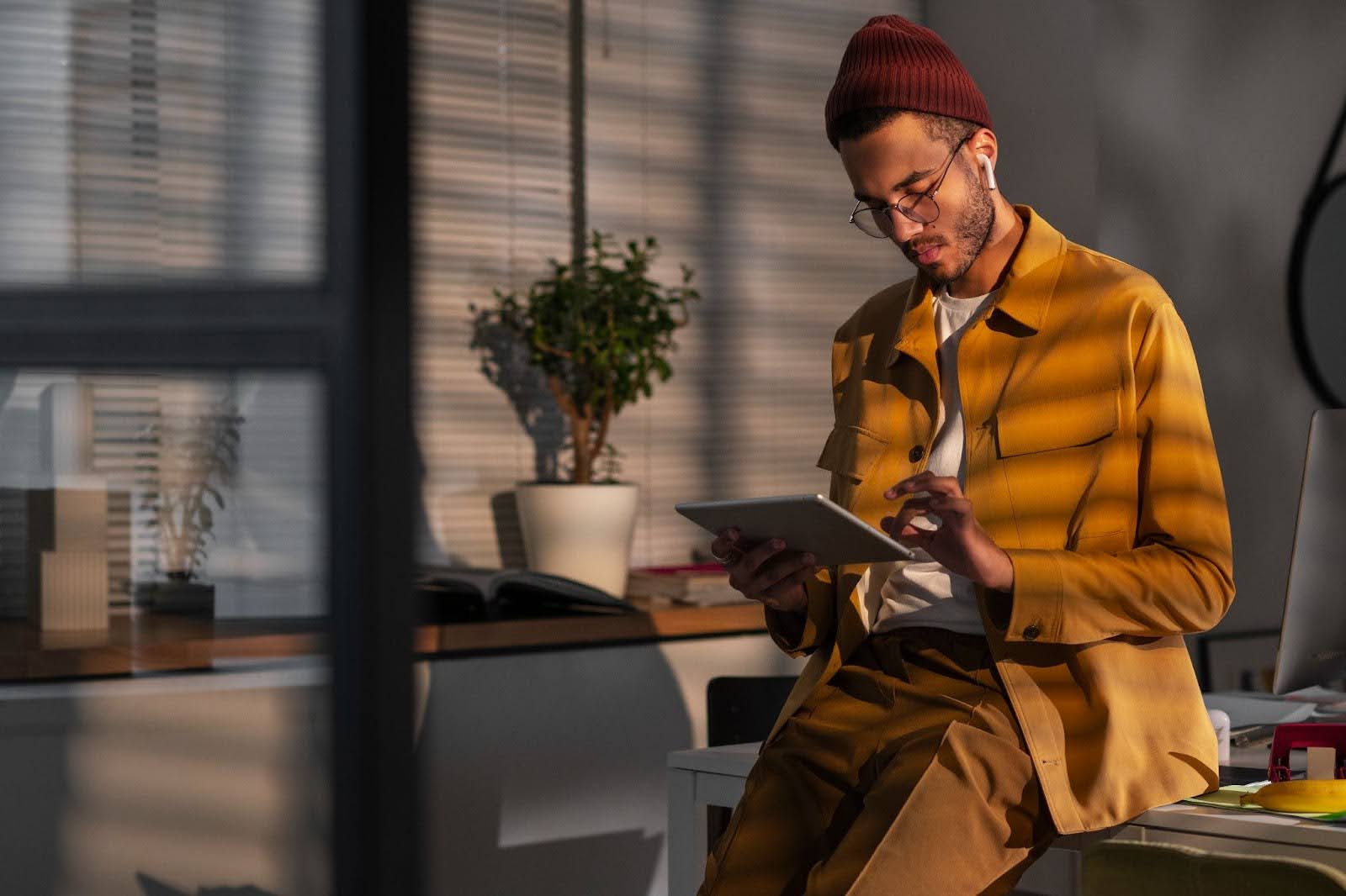 A imagem mostra um homem jovem em um ambiente de trabalho moderno e estiloso. Ele está usando um conjunto de roupas em tons terrosos (jaqueta e calça mostarda), gorro vermelho escuro e óculos, além de fones de ouvido sem fio. Está concentrado, interagindo com um tablet que segura com uma das mãos enquanto utiliza a outra para tocar a tela.