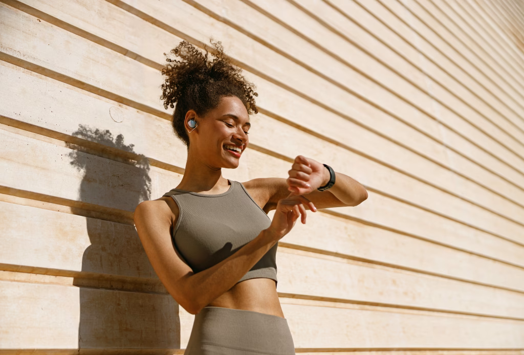 Woman wearing sportswear and earbuds smiling while checking her smartwatch outdoors.